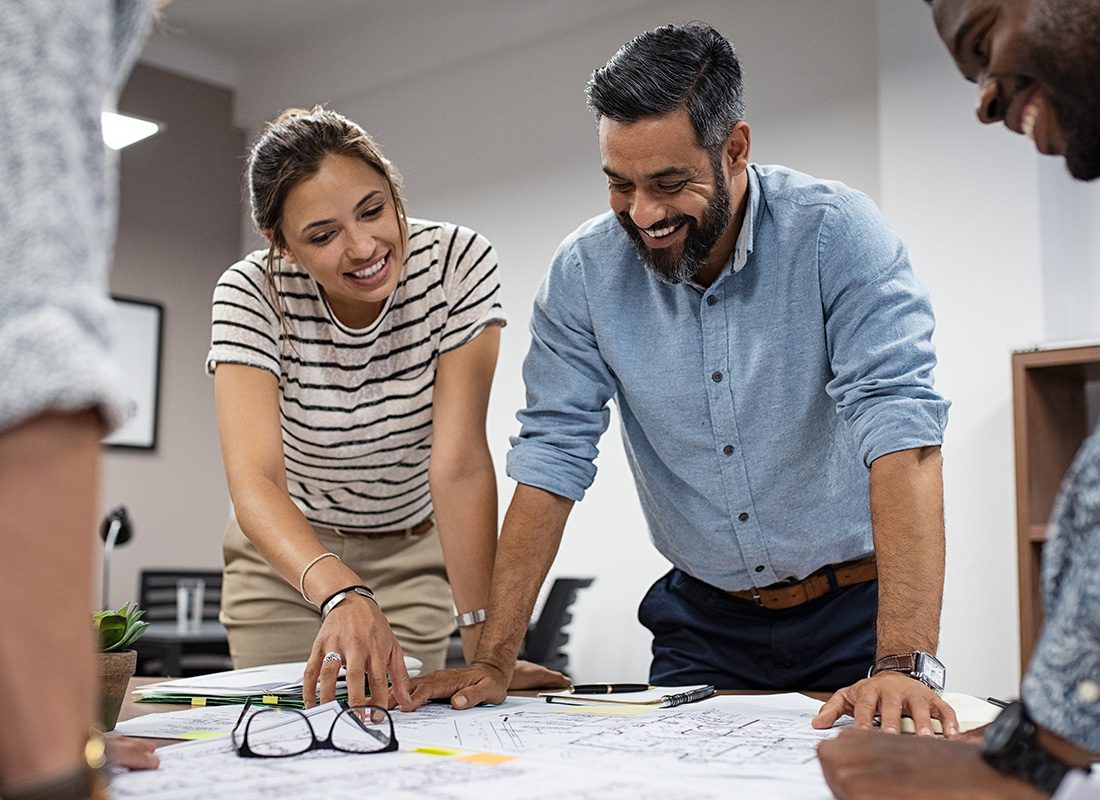 About Our Agency - Portrait of Two Diverse Smiling Architects Looking at Building Blueprints During a Meeting in the Office