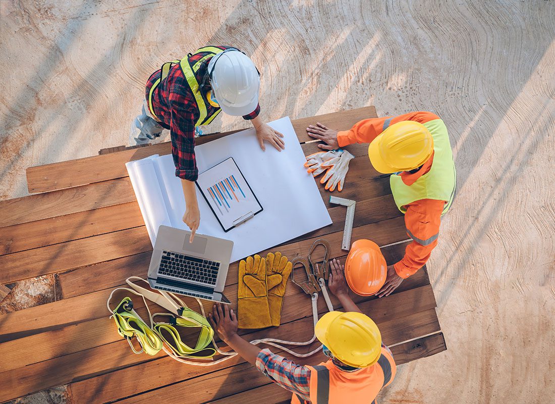 Insurance by Industry - View from Above of Three Contractors Standing Around a Table Outside Planning Out a Construction Project Using Blueprints and a Laptop